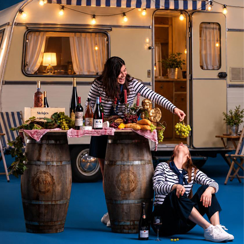 De Meisjes van de Wijn bij een sfeervol verlichte caravan met verschillende flessen wijn, druiven en wijnvaten.